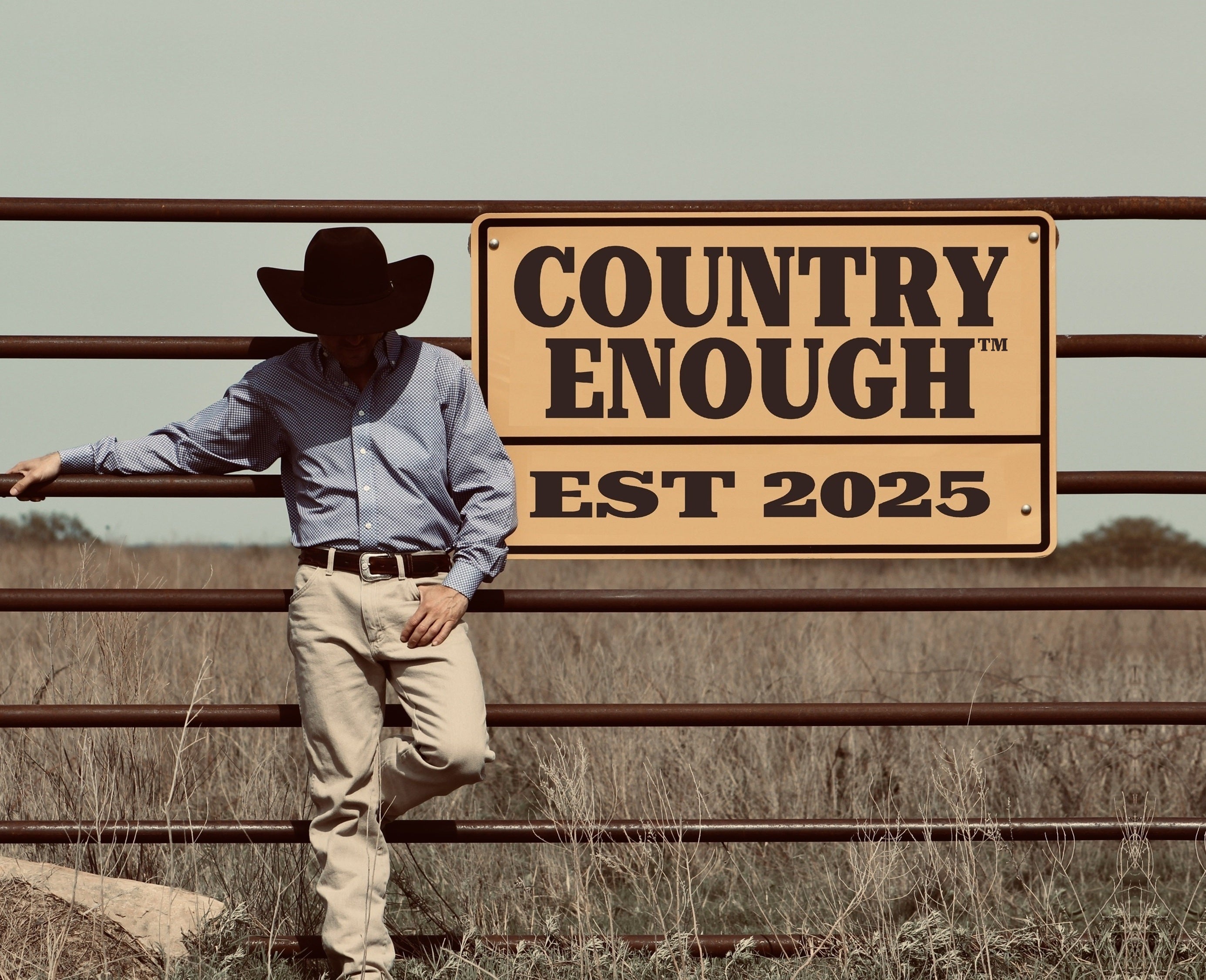 The owner of Country Enough wearing a cowboy, hat leaning against a fence with 'Country Enough' EST 2025 sign in a rural setting.