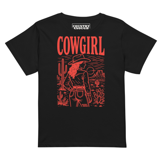 Black t-shirt of cowgirl standing in a desert scene with words cowgirl. all in bright red coloring #black