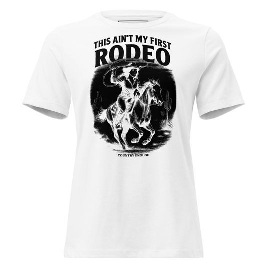 White t-shirt with graphic of a cowgirl on a horse, lassoing, in front of a desert landscape with words this ain't my first rodeo above #white